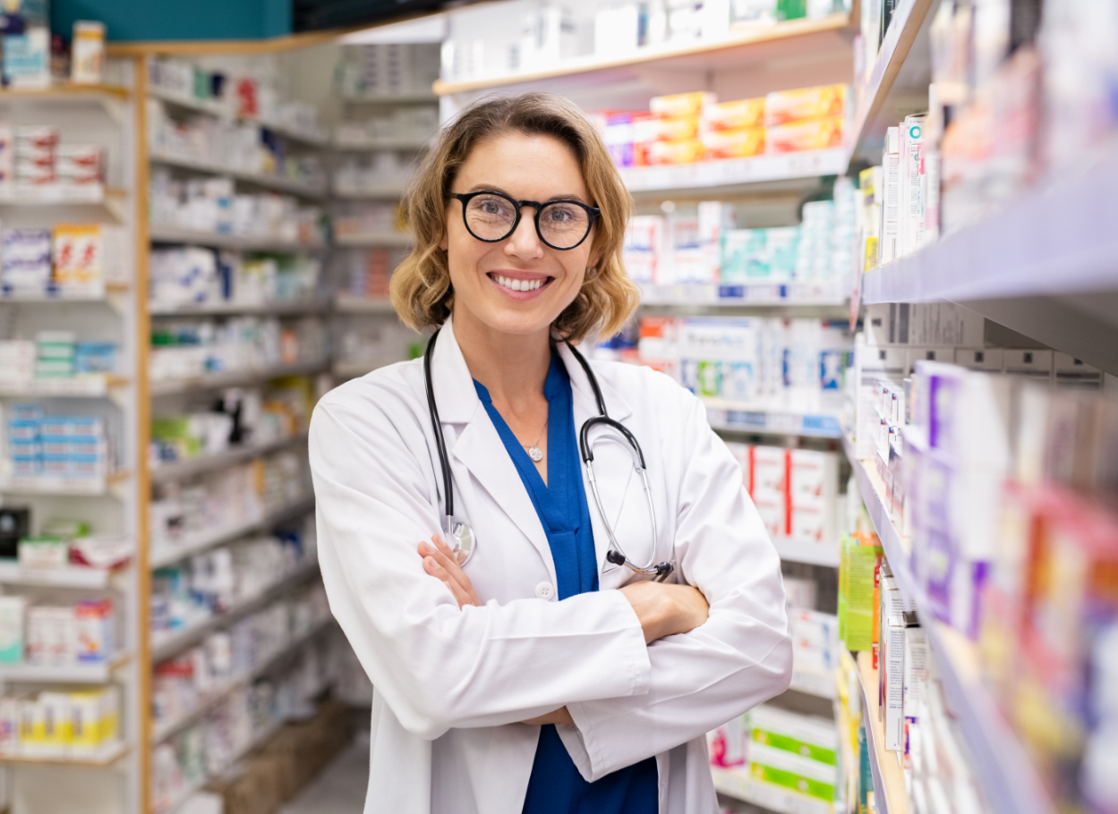 Pharmacist smiling in a pharmacy with a display of medication, representing advocacy and independent pharmacy support in Alberta.